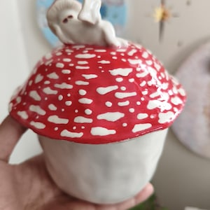 Peut inclure: Un récipient en céramique en forme de champignon, avec un chapeau rouge parsemé de taches blanches. Une petite figurine blanche est perchée au sommet. Le récipient a une base blanche et est tenu dans une main.