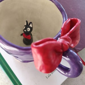 Könnte beinhalten: Eine lila Keramiktasse mit einem roten schleifenförmigen Griff. In der Tasse befindet sich eine kleine schwarze Katzenfigur mit roter Fliege. Die Tasse steht auf einem Buch mit dem Text "Lala Bona".
