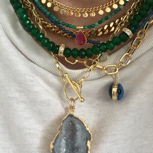 May include: A layered gold chain necklace with a large, gold-rimmed, grey agate pendant. The necklace also features a smaller, red teardrop pendant and a blue evil eye charm.