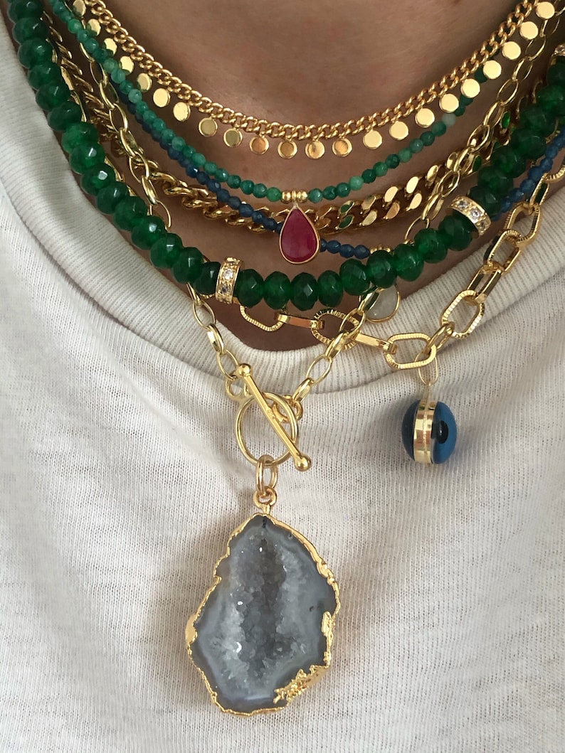 May include: A layered gold chain necklace with a large, gold-rimmed, grey agate pendant. The necklace also features a smaller, red teardrop pendant and a blue evil eye charm.