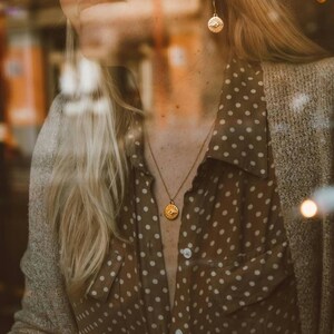 Puede incluir: Una mujer lleva una camisa marr&oacute;n con lunares y un collar y pendientes de oro con un dise&ntilde;o de ojo. El collar y los pendientes son de oro y tienen un dise&ntilde;o simple y elegante.