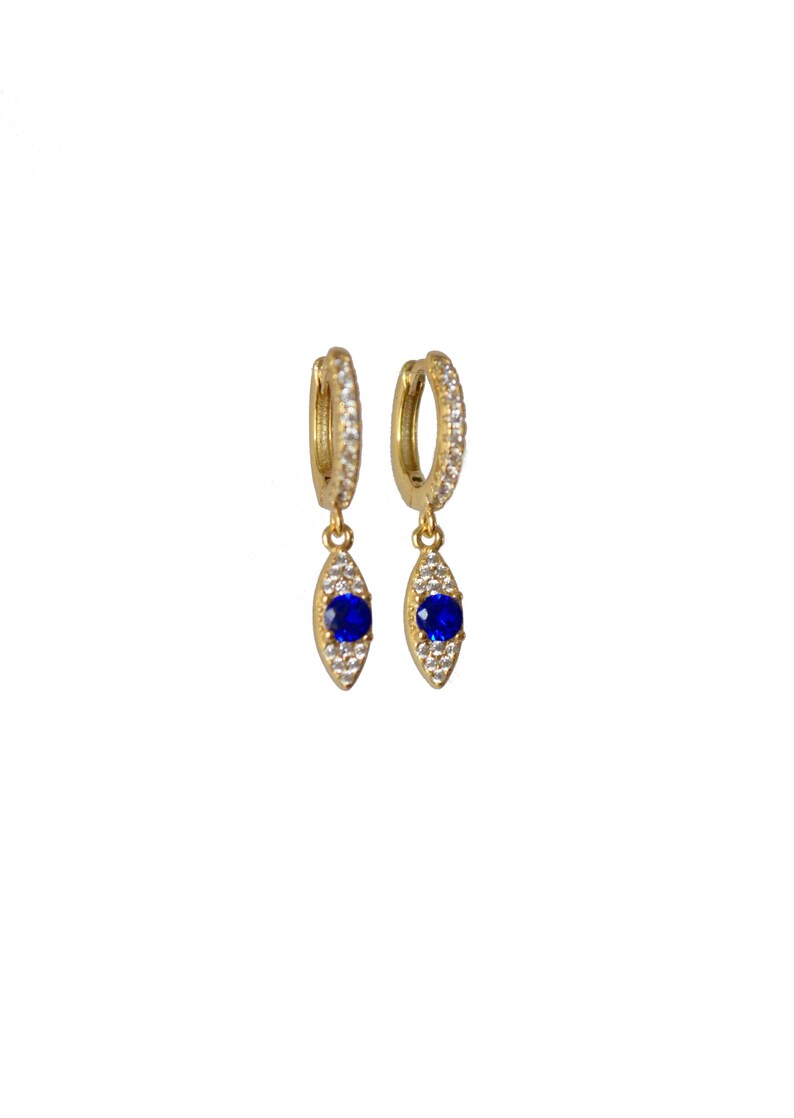 May include: A pair of gold-coloured hoop earrings, each adorned with small, clear crystals. Each earring features a teardrop-shaped pendant with a vibrant blue gemstone at its centre, encircled by smaller, clear crystals. The earrings are set against a white backdrop.