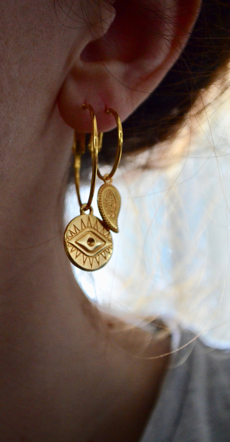Puede incluir: Aretes de aro de oro con un peque&ntilde;o dije de oro colgando de uno de los aros. El dije es un c&iacute;rculo con un dise&ntilde;o de ojo estilizado y un dije en forma de paisley.