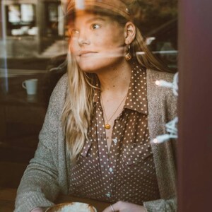 Puede incluir: Una mujer con un c&aacute;rdigan marr&oacute;n, una camisa marr&oacute;n de lunares y una gorra de conductor de noticias marr&oacute;n est&aacute; sentada en una mesa con una taza de caf&eacute;. Lleva pendientes de aro dorados y un collar de oro con un colgante.
