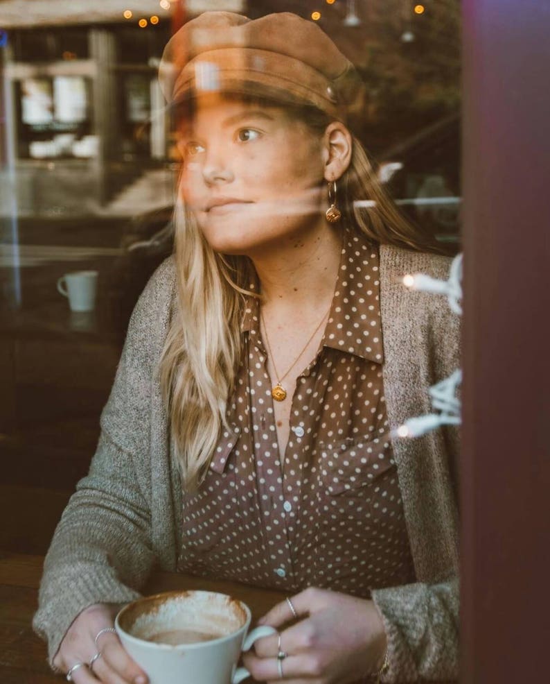 Puede incluir: Una mujer con un c&aacute;rdigan marr&oacute;n, una camisa marr&oacute;n de lunares y una gorra de conductor de noticias marr&oacute;n est&aacute; sentada en una mesa con una taza de caf&eacute;. Lleva pendientes de aro dorados y un collar de oro con un colgante.