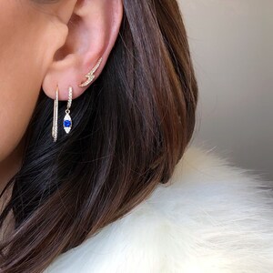 May include: Close-up of three gold earrings on an ear. One is a small lightning bolt shape, another is a hoop with a blue gem, and the third is a long, thin, straight earring. The earrings are adorned with small, sparkling stones.