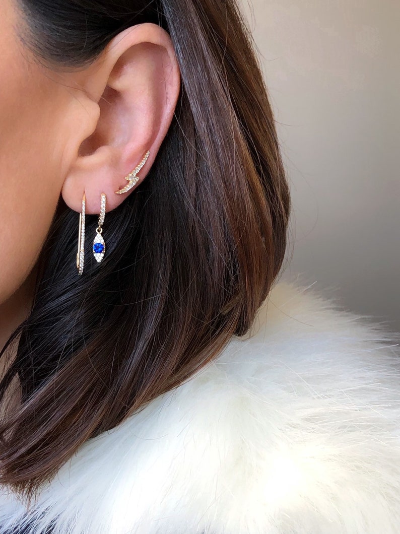May include: Close-up of three gold earrings on an ear. One is a small lightning bolt shape, another is a hoop with a blue gem, and the third is a long, thin, straight earring. The earrings are adorned with small, sparkling stones.