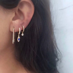 May include: A selection of gold earrings. One is a long, thin bar with small, clear stones. Another is a small hoop with clear stones, and a third has a blue teardrop-shaped stone and a small clear stone.