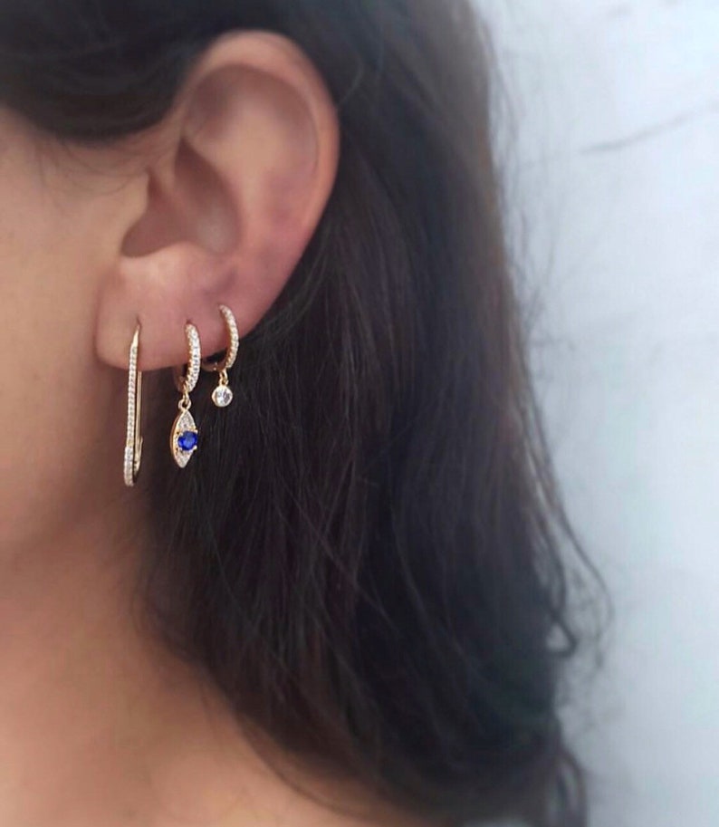 May include: A selection of gold earrings. One is a long, thin bar with small, clear stones. Another is a small hoop with clear stones, and a third has a blue teardrop-shaped stone and a small clear stone.