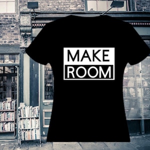 Może przedstawiać: Czarny t-shirt z napisem "MAKE ROOM" w białym prostokącie. Koszulka jest prezentowana na tle księgarni z półkami na książki i ceglanym budynkiem.