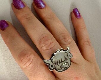 May include: A silver ring featuring a cartoon-style Highland cow head. The ring is worn on a person's finger.