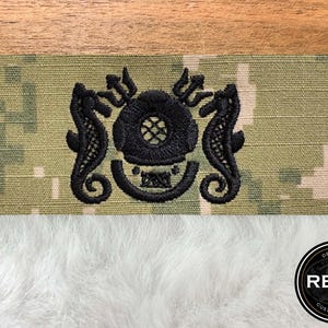 May include: A black embroidered patch featuring a diving helmet, seahorses, and a trident on a camouflage background. The patch has the text "REV 7 CUSTOM MADE" in a circular logo. The patch is on a white fluffy surface.