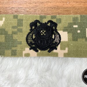 May include: A black embroidered diving helmet patch is centered on a camouflage fabric strip. The patch is detailed with intricate lines and shapes. The background is a digital camouflage pattern in shades of green and tan. The bottom of the image shows white fluffy material.