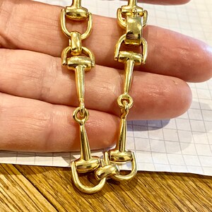 Pode incluir: Um par de brincos dourados com um design de freio de cavalo. Os brincos t&ecirc;m uma forma longa e esguia com uma s&eacute;rie de elos conectados e um freio detalhado na parte inferior. Os brincos s&atilde;o mostrados em um fundo neutro.
