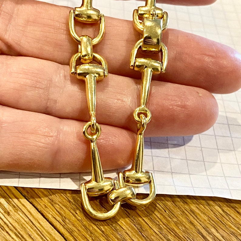 Pode incluir: Um par de brincos dourados com um design de freio de cavalo. Os brincos t&ecirc;m uma forma longa e esguia com uma s&eacute;rie de elos conectados e um freio detalhado na parte inferior. Os brincos s&atilde;o mostrados em um fundo neutro.