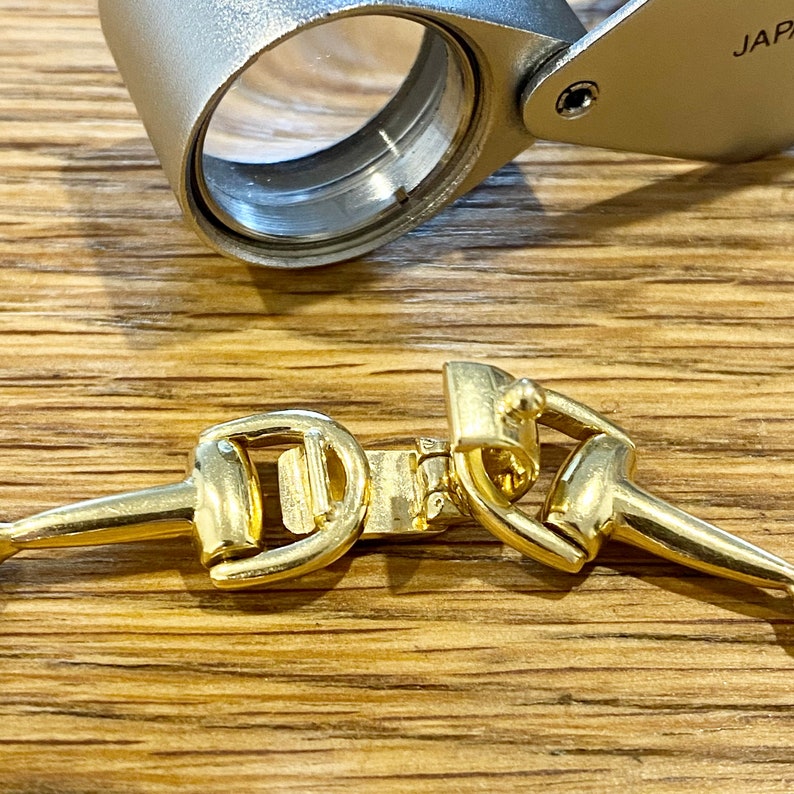 Pode incluir: Close-up de um fecho de pulseira de metal dourado em estilo equestre. O fecho tem a forma de um freio de cavalo. Uma lupa prateada est&aacute; no fundo.