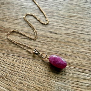May include: A delicate gold-tone chain necklace featuring a single, smooth, oval-shaped magenta gemstone pendant. The pendant is securely attached to the chain with a small metal bail, showcasing a vibrant pink hue and a polished, reflective surface.