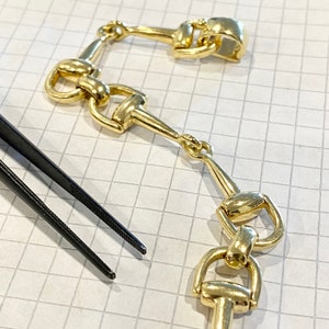 Pode incluir: Close-up de elos de corrente dourados em estilo equestre, possivelmente para joias ou um elemento decorativo. Os elos est&atilde;o dispostos sobre uma superf&iacute;cie quadriculada, com uma pin&ccedil;a preta no canto inferior esquerdo.