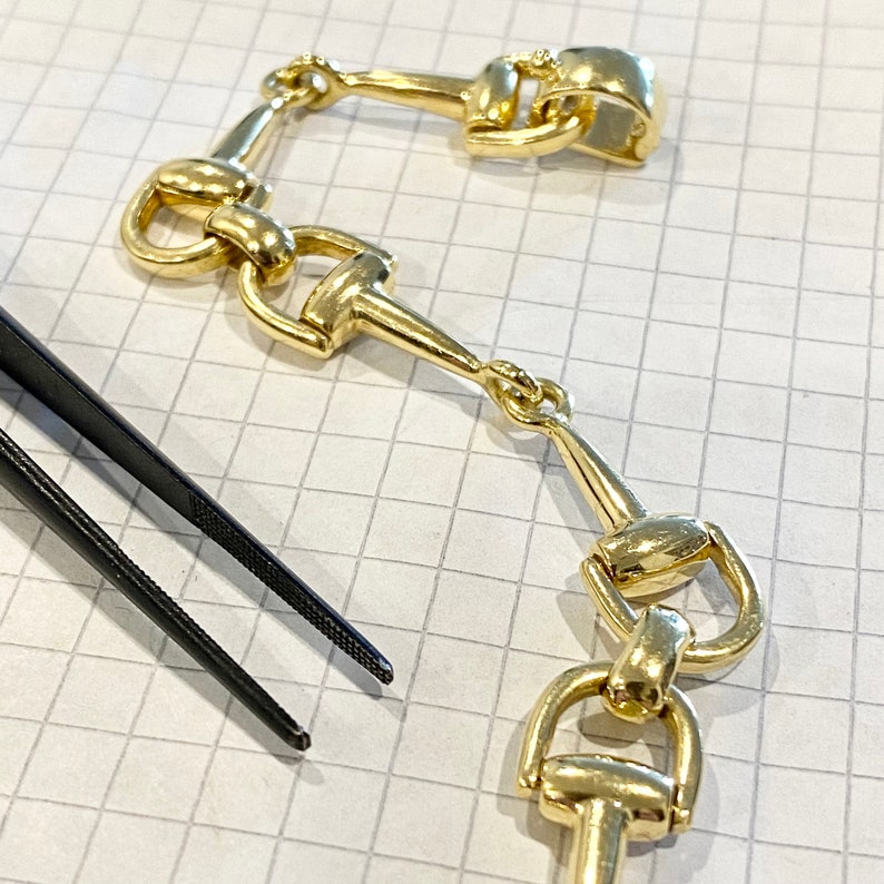 Pode incluir: Close-up de elos de corrente dourados em estilo equestre, possivelmente para joias ou um elemento decorativo. Os elos est&atilde;o dispostos sobre uma superf&iacute;cie quadriculada, com uma pin&ccedil;a preta no canto inferior esquerdo.