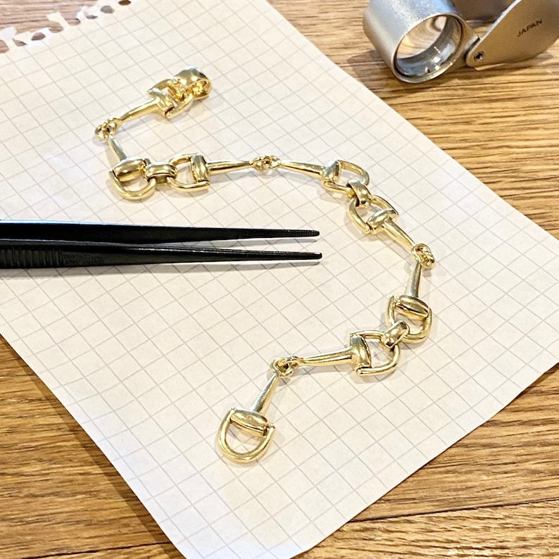 Pode incluir: Uma pulseira dourada com uma s&eacute;rie de elos conectados em forma de freio de cavalo. A pulseira &eacute; exibida em papel quadriculado branco, com um par de pin&ccedil;as pretas e uma lupa no fundo.