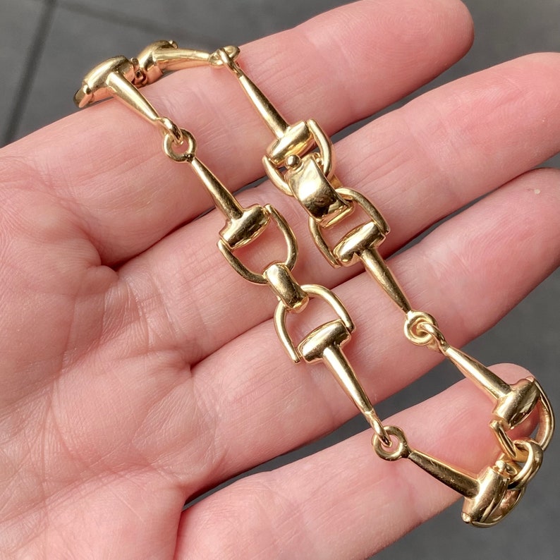 Pode incluir: Uma pulseira dourada com um design de elos que lembra equipamentos equestres. A pulseira tem uma s&eacute;rie de elos conectados, em forma de freio, criando um acess&oacute;rio &uacute;nico e elegante. O metal tem um acabamento polido, refletindo a luz.