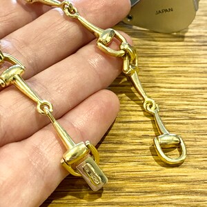 Pode incluir: Um colar dourado com um design de freio de cavalo. O colar tem uma s&eacute;rie de elos conectados em forma de freio, com um fecho na extremidade. A joia &eacute; exibida em uma superf&iacute;cie de madeira.