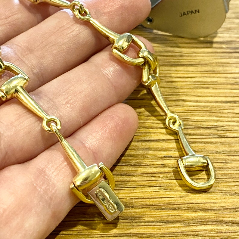 Pode incluir: Um colar dourado com um design de freio de cavalo. O colar tem uma s&eacute;rie de elos conectados em forma de freio, com um fecho na extremidade. A joia &eacute; exibida em uma superf&iacute;cie de madeira.