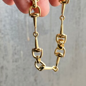 Pode incluir: Close-up de um colar de corrente dourada com um design de freio de cavalo. O colar &eacute; feito de v&aacute;rios elos conectados, cada um com uma forma de freio detalhada. Os elos s&atilde;o conectados por pequenos an&eacute;is circulares. A joia &eacute; exibida em um fundo cinza.