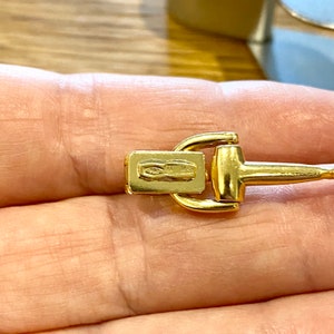 Pode incluir: Close-up de um fecho de colar dourado. O fecho apresenta uma barra retangular com marca&ccedil;&otilde;es estampadas e um design em forma de estribo. O metal tem um acabamento polido, refletindo a luz.