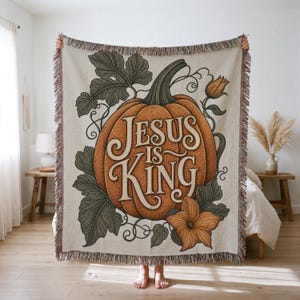 Könnte beinhalten: Eine gewebte Wohndecke mit einem großen orangefarbenen Kürbis, grünen Blättern und Ranken. Der Text 'JESUS IS KING' ist in einer dekorativen Schriftart in der Mitte des Kürbisses abgebildet. Die Decke hat einen cremefarbenen Hintergrund und Fransen.