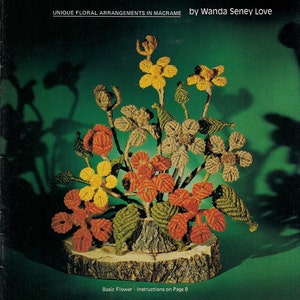 May include: A book cover with a green background and the title "BLOOMIN' MACRAME" in yellow and green letters. The book cover features a macrame flower arrangement with orange, yellow, and brown flowers on a wooden base. The text "UNIQUE FLORAL ARRANGEMENTS IN MACRAME by Wanda Seney Love" is printed below the title.