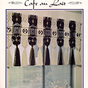 Op de afbeelding: Een macramé-raambekleding met de titel "Cafe au Lait". De bekleding is gemaakt van bruin en wit garen en heeft een franje aan de onderkant. De bekleding hangt aan een raam met witte jaloezieën.