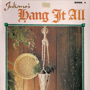 May include: A macrame plant hanger with a white cord and a wooden bead at the top. The hanger is hanging from a wooden wall and has a green plant inside. The text "Hang It All" is written in a decorative font above the hanger.
