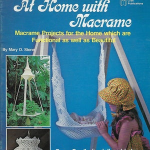 Peut inclure: Une couverture de livre avec le titre "At Home with Macrame" et le sous-titre "Macrame Projects for the Home which are Functional as well as Beautiful". La couverture présente un porte-plante suspendu en macramé blanc, un berceau en macramé blanc et une fille tenant une poupée dans le berceau. Le livre est de Mary O. Stone et est publié par Craft Publications.