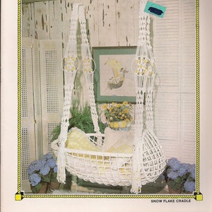 Könnte beinhalten: Eine weiße Makramee-Babywiege, die von der Decke hängt. Die Wiege ist aus weißem Seil gefertigt und hat eine weiße Stoffauskleidung. Die Wiege ist mit weißen Perlen verziert. Die Wiege hängt in einem Raum mit weißen Wänden und einem weißen Holzboden. Der Text "Snow Flake Cradle" ist unter dem Bild gedruckt.
