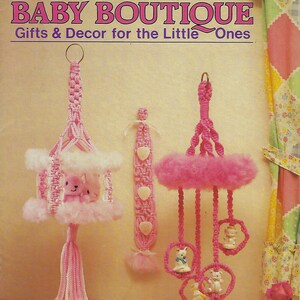 May include: A pink and white crocheted baby mobile with three hanging pink rings and white stuffed animals. The mobile is hanging from a pink ceiling hook. The image also shows a pink and white crocheted crib canopy with a white stuffed animal inside. The image has the text "BABY BOUTIQUE Gifts & Decor for the Little Ones" and "MM 481".