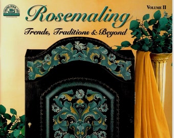 May include: A book titled "Rosemaling: Trends, Traditions & Beyond, Volume II" by Shirley Peterich, CDA and Pam Rucinski, VGM. The cover features a black painted wooden cabinet with a green, gold, and white floral design. The cabinet is on a table with a teal surface. There are two smaller black painted wooden plates with similar floral designs on the table.