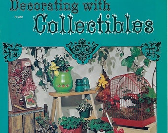 以下が含まれることがあります： ターコイズブルーの背景に黒い文字で「Decorating with Collectibles」というタイトルが書かれた、ビンテージな本の表紙。表紙には、鳥かご、じょうろ、ピッチャー、鉢植えの植物など、さまざまな家庭用装飾品が置かれた静物画が描かれている。