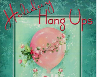 May include: A pink hot air balloon with a Santa Claus figure inside. The balloon is decorated with holly, bells, and candy canes. The text "Holiday Hang Ups" is written in red cursive at the top of the image. The text "By Sandra Malone" is written at the bottom of the image.