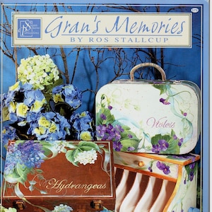 Puede incluir: Un libro titulado "Gran's Memories" de Ros Stallcup, que presenta cajas de madera pintadas con diseños florales. Las cajas están decoradas con hortensias, violetas y otras flores. La portada del libro es azul con un borde blanco.