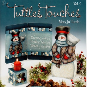 May include: A book cover for "Tuttles Touches" volume 5 by Mary Jo Tuttle. The cover features a painted snowman with a red scarf and plaid shirt holding a shovel. The snowman is standing in front of a blue and white painted wooden box with the text "Snowy Days Frosty Nights Warm Hearts".