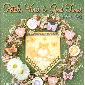 May include: A book titled "Petals, Vines & Good Times" by Laurie Speltz, featuring a wreath with flowers, butterflies, and hearts. The book cover also includes a painted sign with a cat and flowers.
