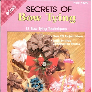 May include: A book titled "Secrets of Bow Tying" with a pink cover and a plaid pattern on the top and bottom. The book features a photo of various gift baskets with bows tied in different styles. The book cover also includes the text "13 Bow Tying Techniques", "Over 20 Project Ideas", and "Step-By-Step Construction Photos".