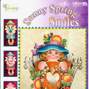 Può includere: Un portacandele votivo con un'apertura a forma di cuore e un design fantasioso con una ragazza con un cappello blu e fiori. Il portacandele è in legno e dipinto con colori vivaci. Il testo "Sunny Spring Smiles" è stampato nella parte superiore dell'immagine.