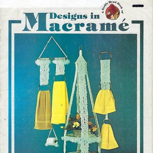 Può includere: Una copertina di libro con il titolo "Designs in Macrame" e un'immagine di un portavasi in macramè bianco con due ripiani e un portasciugamani in macramè bianco con due asciugamani.