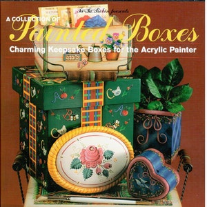 May include: A collection of painted wooden boxes featuring various designs, including a heart-shaped box, a round box with a floral design, and a box with a plaid pattern. The boxes are stacked on top of each other and have a rustic, charming look. The title of the book is "Painted Boxes: Charming Keepsake Boxes for the Acrylic Painter" by Ta Ta Robin.