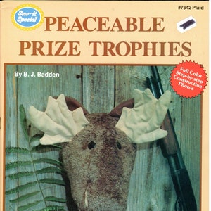Puede incluir: Una portada de libro con un trofeo de cabeza de alce marrón sobre un fondo de madera. El título del libro es "Peaceable Prize Trophies" y el subtítulo es "Four Sportsman-Look Soft Sculpture Trophies". La portada del libro también incluye el texto "Full Color Step-by-step Construction Photos" y "Full-Size Patterns and Complete Instructions".