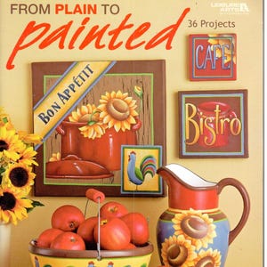 Könnte beinhalten: Buchcover "FROM PLAIN TO painted" mit dekorativer Malerei. Das Bild zeigt einen roten Krug mit Sonnenblumenmotiven, einen gelben Korb mit Hahnmotiven und ein Gemälde eines Topfes mit Sonnenblumen. Der Text "BON APPÉTIT" ist sichtbar.