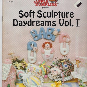 May include: A book titled "Soft Sculpture Daydreams Vol. I" by Apple Dumplins. The book cover features a pink background with white polka dots and a variety of handmade baby-themed items, including a baby rattle, a tooth fairy pillow, and a pincushion.