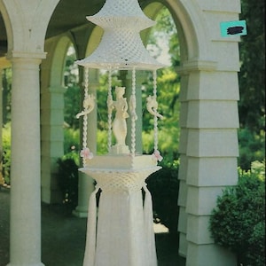 May include: A white macrame fountain, "Fountain of the Orient," suspended from a porch. The fountain features a tiered, pagoda-style top, a central statue, and hanging ornaments. The design includes tassels and pink floral accents, with the title "Macrame Enchantment" at the top.